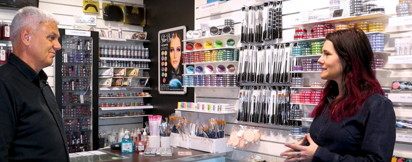 Interviewer and business owner standing in her cosmetics store, mid shot of their profiles as they stand facing one another.